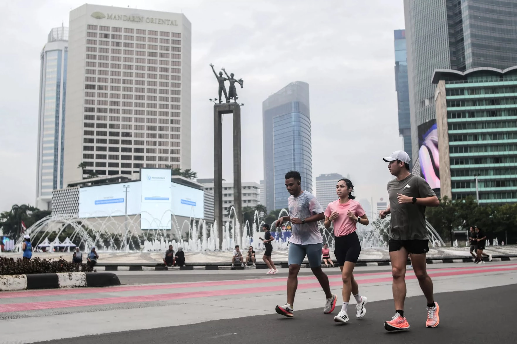 Tiga orang berlari di kawasan Bundaran HI Jakarta dengan latar gedung dan suasan kota